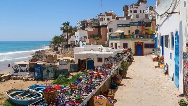A coastal street with buildings, a beach, and a row of colorful items on display. - Duplex feet in water in Taghazout 4563 (Agadir)