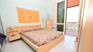 Une chambre avec un lit en bois, une table de chevet, un ventilateur fixé au mur et une fenêtre donnant sur l’extérieur.