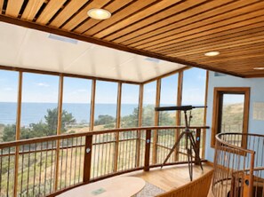 A room with a large window offering a view of the sea, a wooden ceiling, and a telescope on a stand.