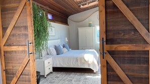 A bedroom with a wooden ceiling, a bed with white bedding, a nightstand, a white wardrobe, and a large window. - KH: Life is a Beach (Dillon Beach)