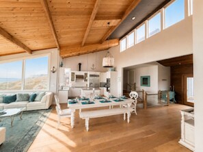 A spacious living area with a wooden ceiling, a dining table with blue placemats, and a view of the outdoors through large windows. - KH: Life is a Beach (Dillon Beach)