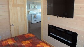 Habitació petita amb un llit, una llar de foc, una televisió i una cuina visible a través d'una porta oberta.