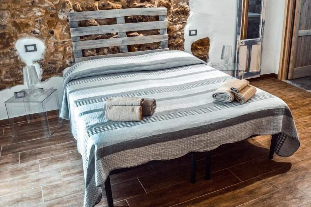 Un lit avec une couvre-lit à rayures et deux serviettes enroulées, une tête de lit en bois et un mur de pierre en arrière-plan.
