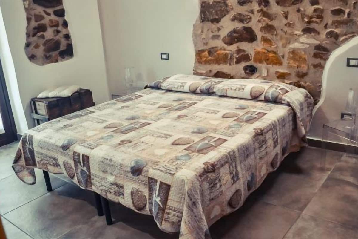 Une chambre avec un mur en pierre, un lit recouvert d’une couvre-lit à motifs, une table de chevet et une table en verre.