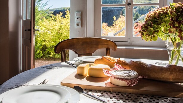 A dining table set with plates, cheese, bread, and a vase with flowers, positioned near a window with a view of greenery and hills. - Would you like to let your mind wander? Welcome to the Morvan Nature Park (Cussy-en-Morvan)