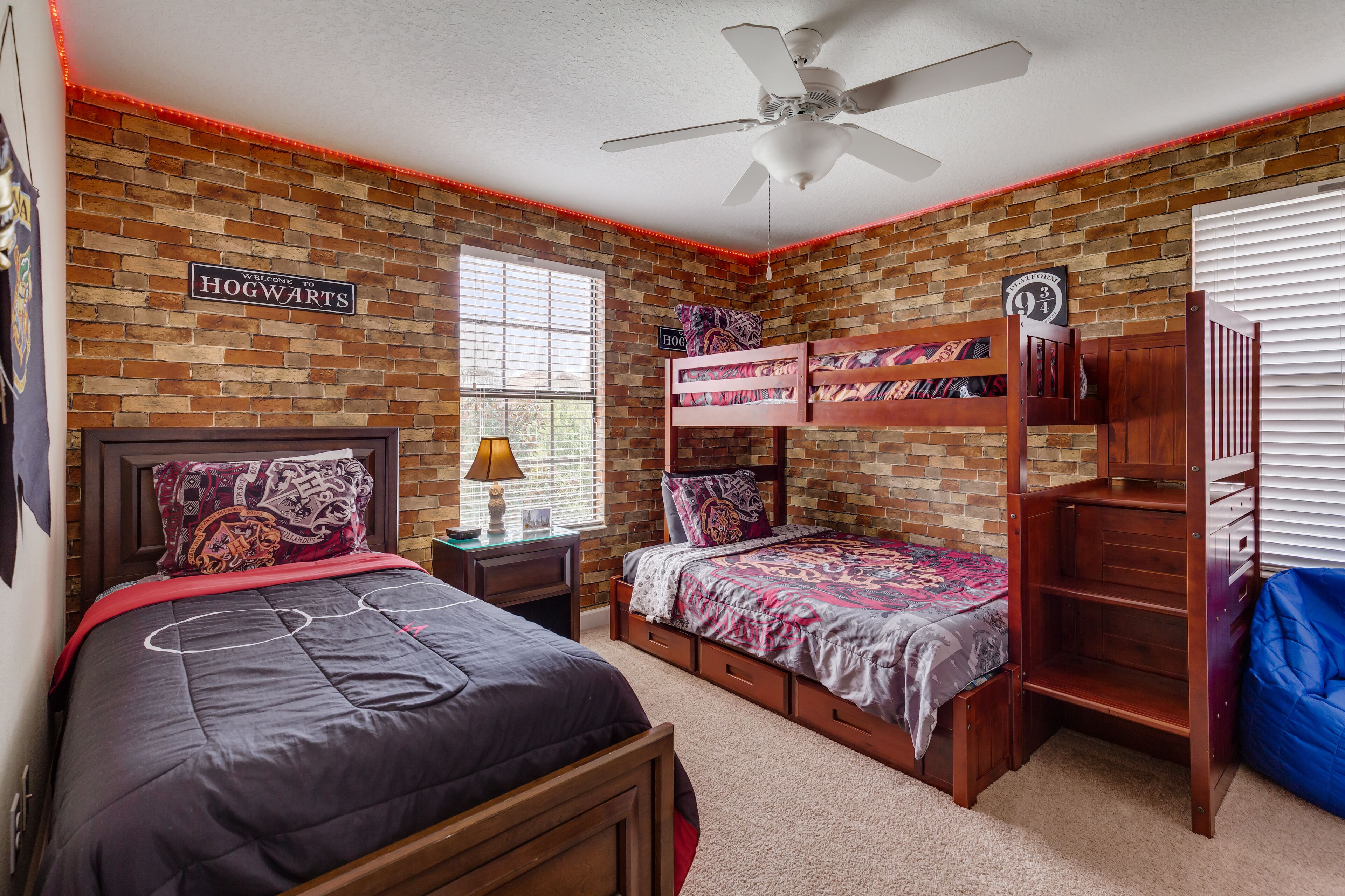 A room with a brick wall, a ceiling fan, bunk beds, a bed with a red blanket, and a sign that says "Welcome to Hogwarts".