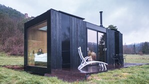 Una cabaña moderna de madera negra con puerta y ventana de vidrio, un pequeño espacio de estar al aire libre y una chimenea.