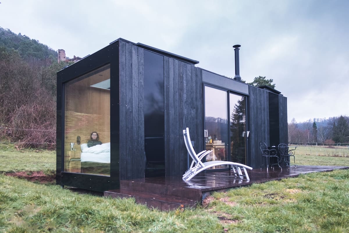 Una cabaña moderna de madera negra con puerta y ventana de vidrio, un pequeño espacio de estar al aire libre y una chimenea.