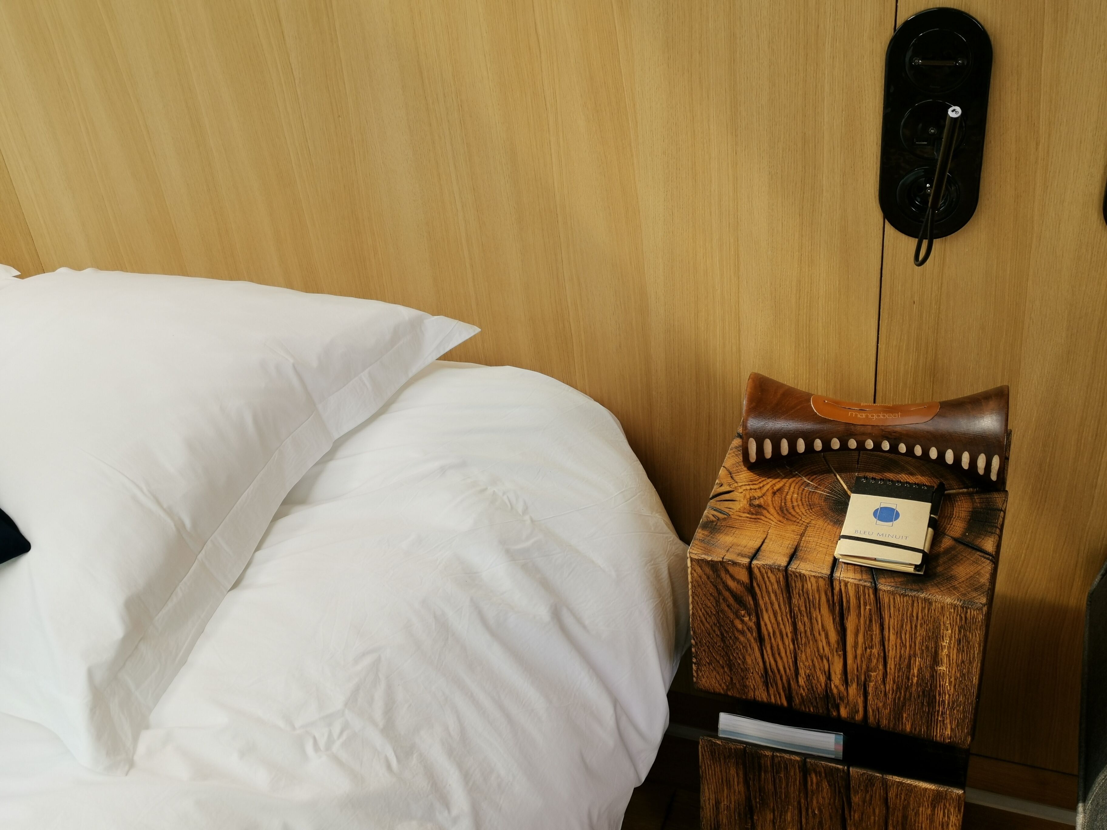 A neatly made bed with white linens, a wooden nightstand with a book and a phone, and a wall-mounted charging station.