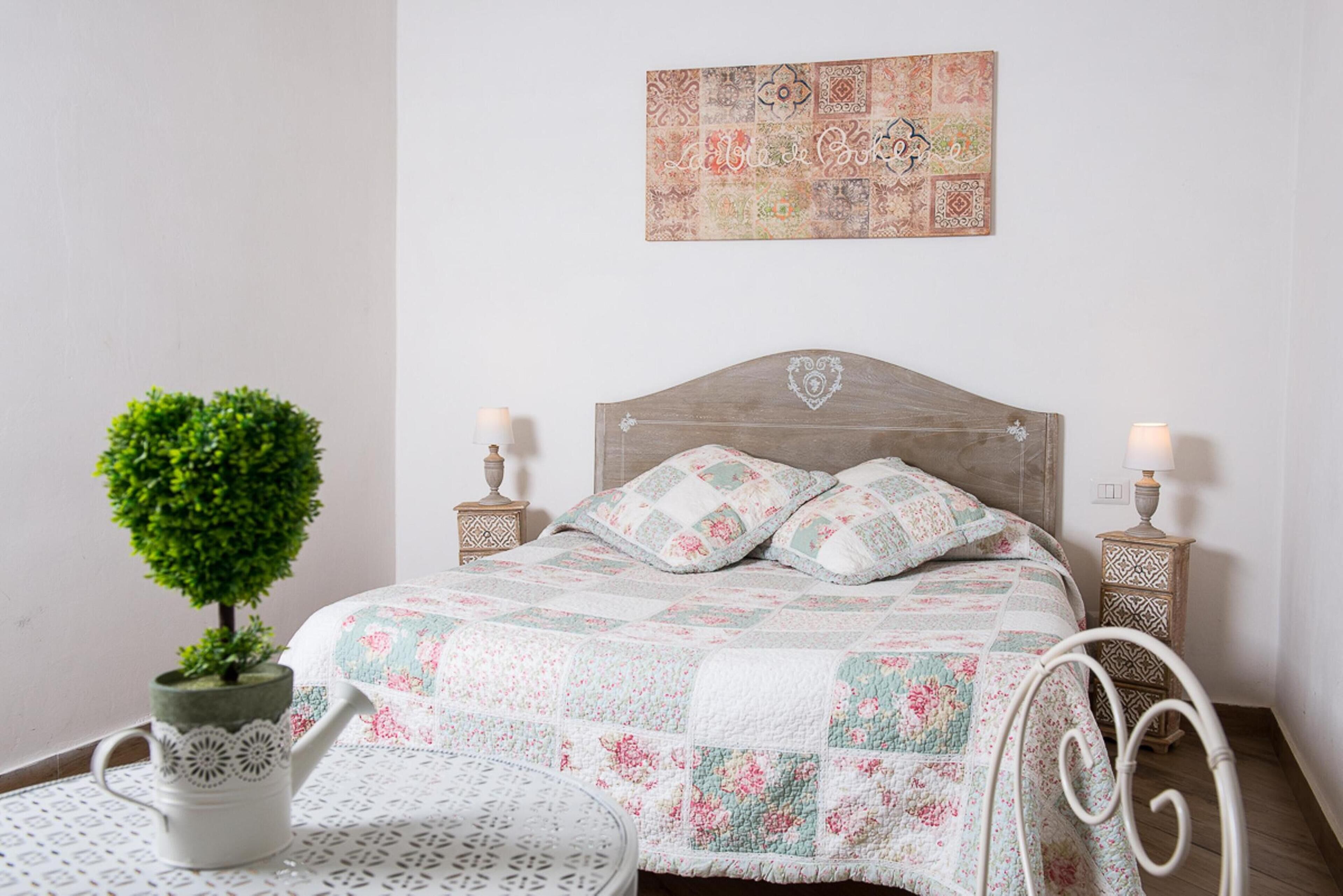A neatly arranged bedroom with a wooden headboard, floral bedding, a small potted plant, and two bedside tables with lamps.