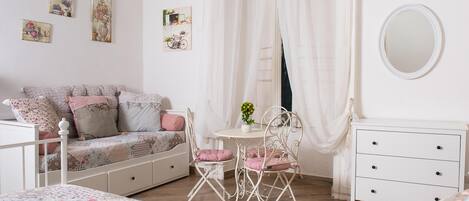 A bedroom with a bed, a white dresser, a chair, a small table with a vase, and a mirror on the wall.