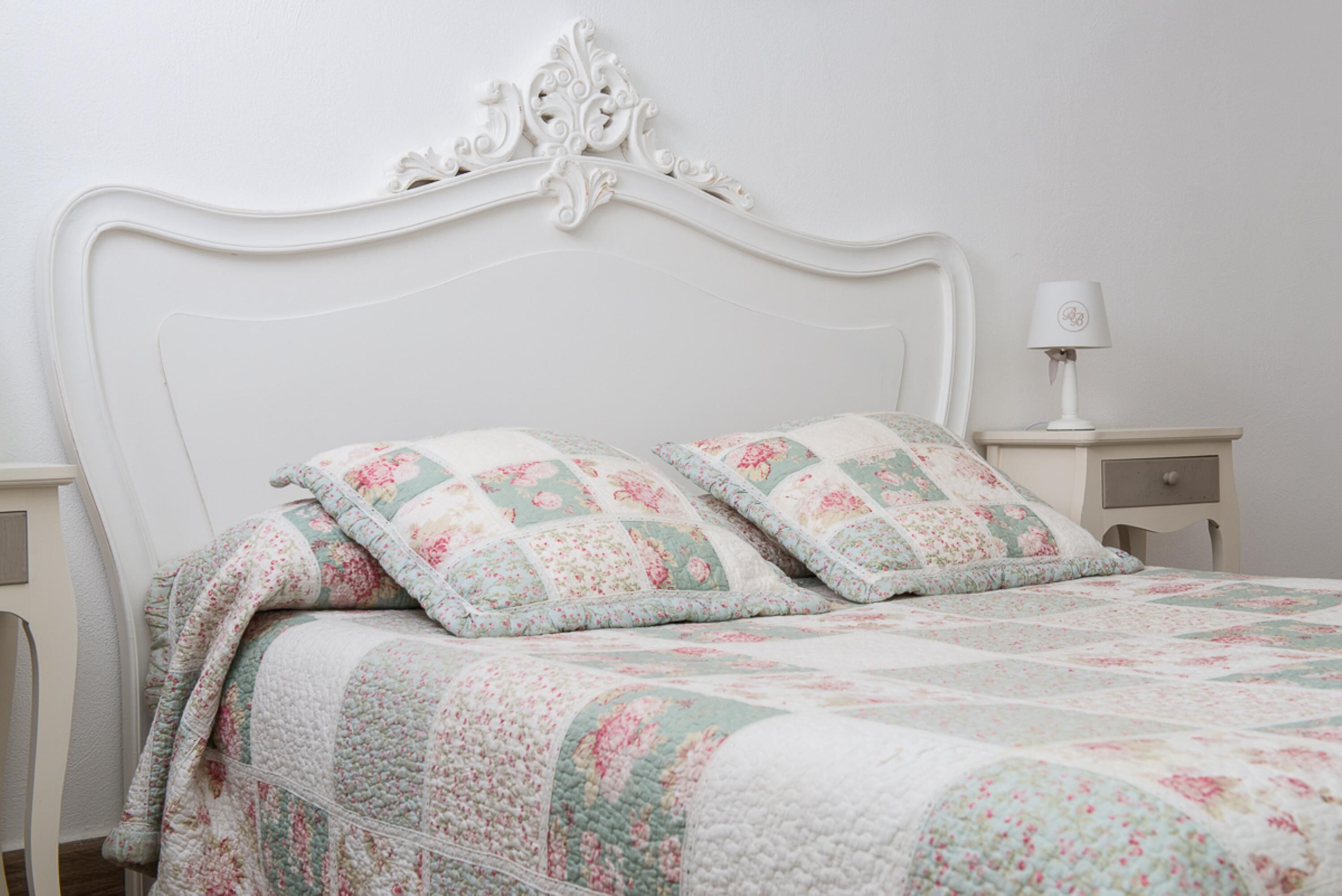 A bed with a floral quilt, a white headboard, and a bedside table with a lamp.