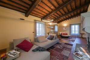 A spacious living room with a high wooden ceiling, a grey sofa with red and grey cushions, a patterned rug, and a dining table with a magazine and a glass of wine. - Design villa with large swimming pool in the heart of the Tuscan countryside (Cortona)