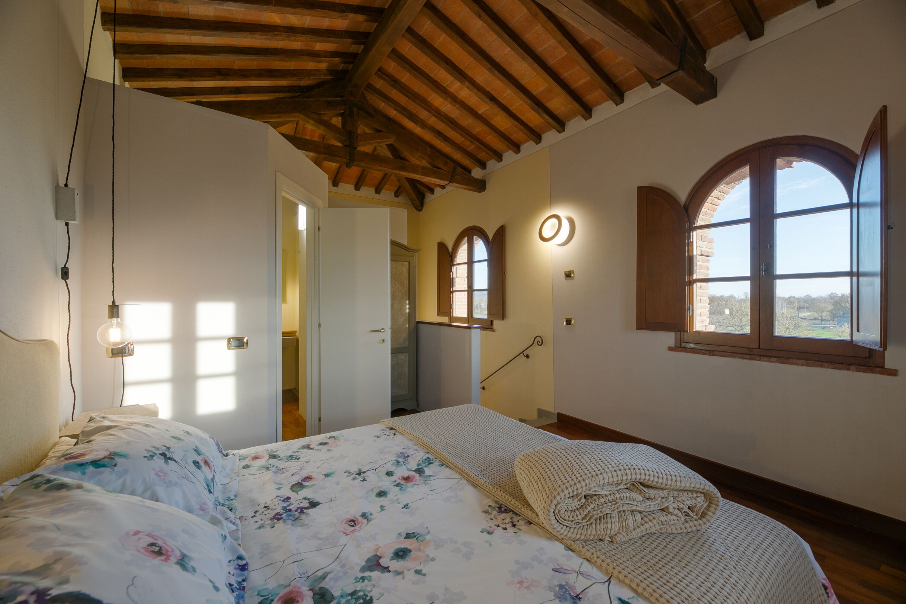 Quarto com teto de madeira, cama com roupa de cama florida, janela com vista e porta para outro cômodo.