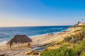 A beach with a thatched hut, rocky shoreline, and a building with palm trees in the background. - Ocean 3 Blocks * Prof Design * AC * Upper Level (La Jolla)