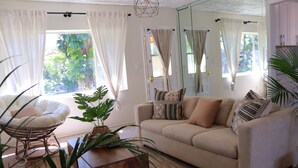 Una sala de estar con un sofá color beige, una mesa de centro de madera y una silla de mimbre. Hay cortinas en las ventanas, y una planta sobre la mesa de centro.