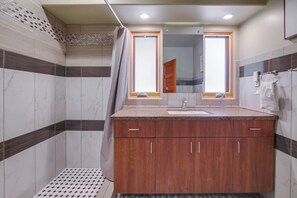 Un baño con tocador de madera, lavamanos y espejo. Cuenta con una zona de ducha con piso de azulejos y cortina.