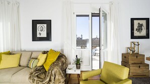 Ein Wohnzimmer mit einer Couch, einem grünen Sessel, einer Katze, einem kleinen Tisch mit einer Pflanze und einer Tür, die auf einen Außenbalkon führt.