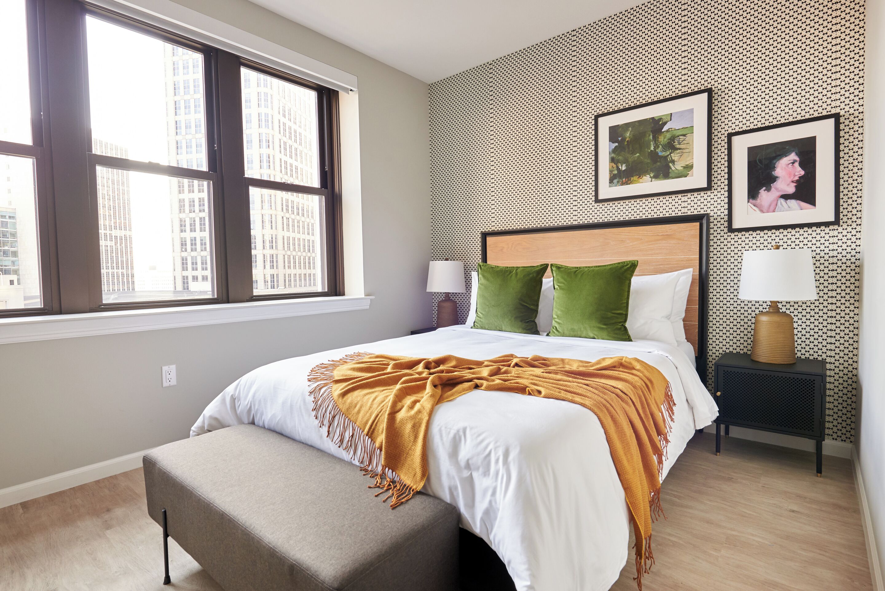 Ein Hotelzimmer mit Bett, Fenster, Nachttisch und einer Wand mit Kunstwerken.