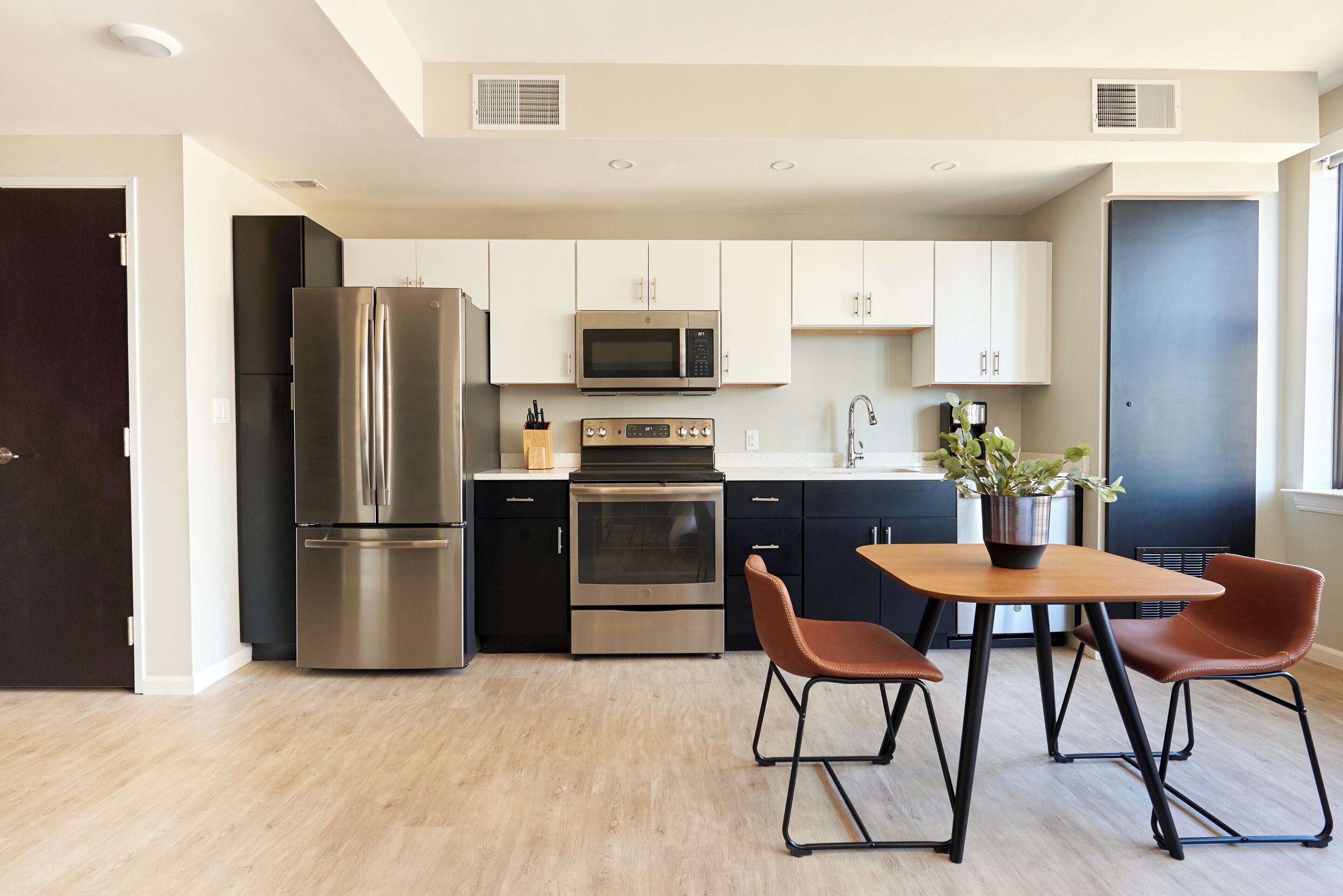 A modern kitchen with stainless steel appliances, a dining area with a wooden table and two chairs, and a dark-colored refrigerator.