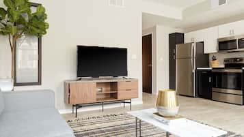 A modern living room with a flat-screen TV, a wooden cabinet, a large plant, and a kitchen area with stainless steel appliances.