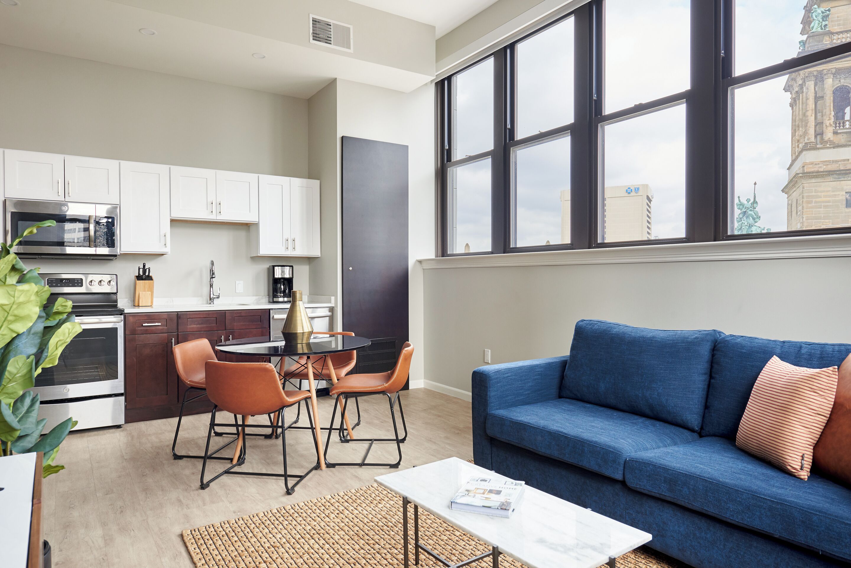A modern kitchen with white cabinets, a dining area with a round table and chairs, and a blue sofa in a living area with large windows.