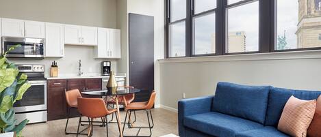 A modern kitchen with white cabinets, a dining area with a round table and chairs, and a blue sofa in a living area with large windows.