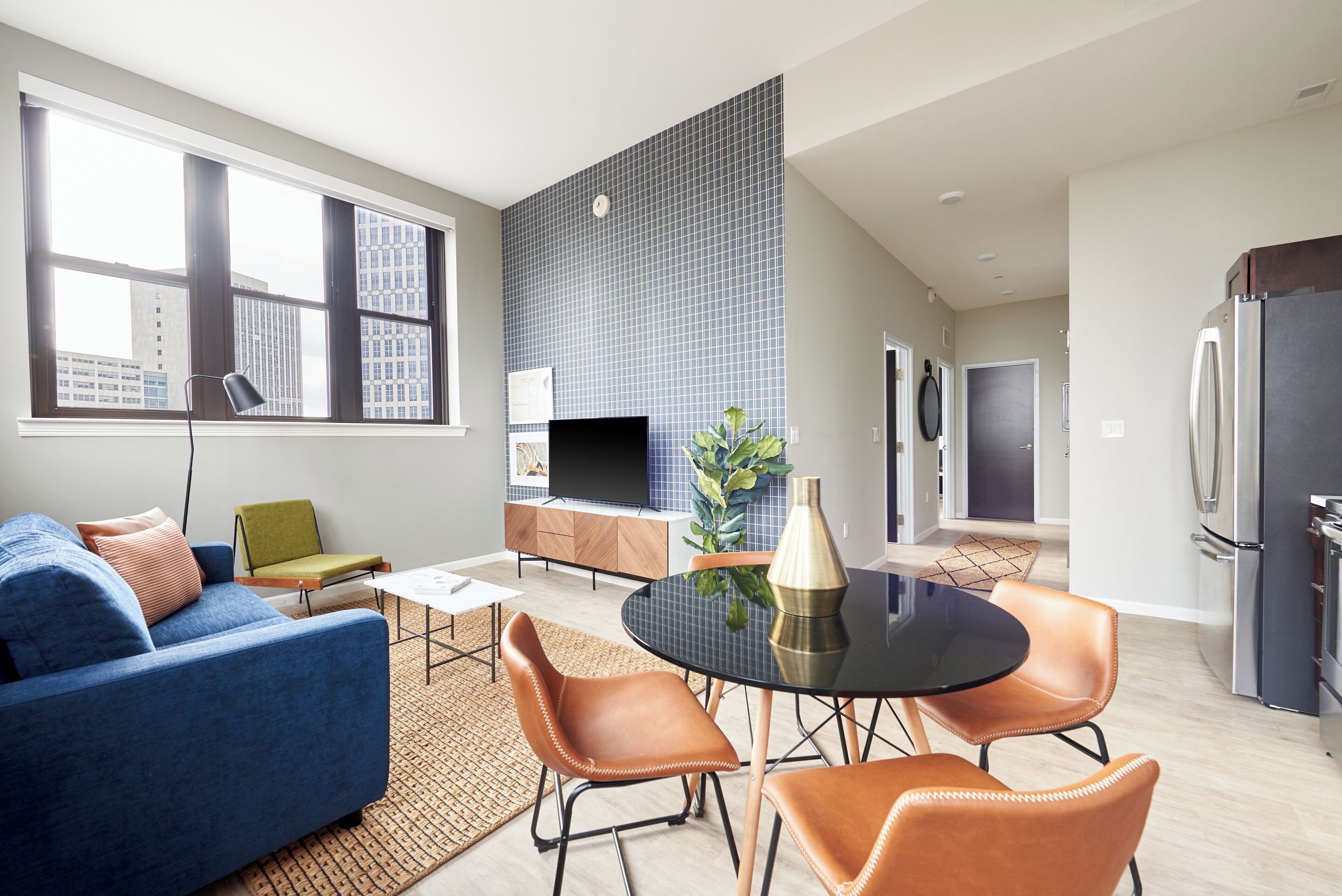A modern living room with a blue sofa, a dining table with chairs, a flat-screen TV, and a kitchen area with a refrigerator.