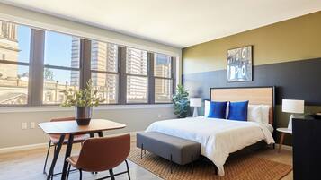 A modern hotel room with a large window, a bed with blue pillows, a small table with chairs, and a potted plant.