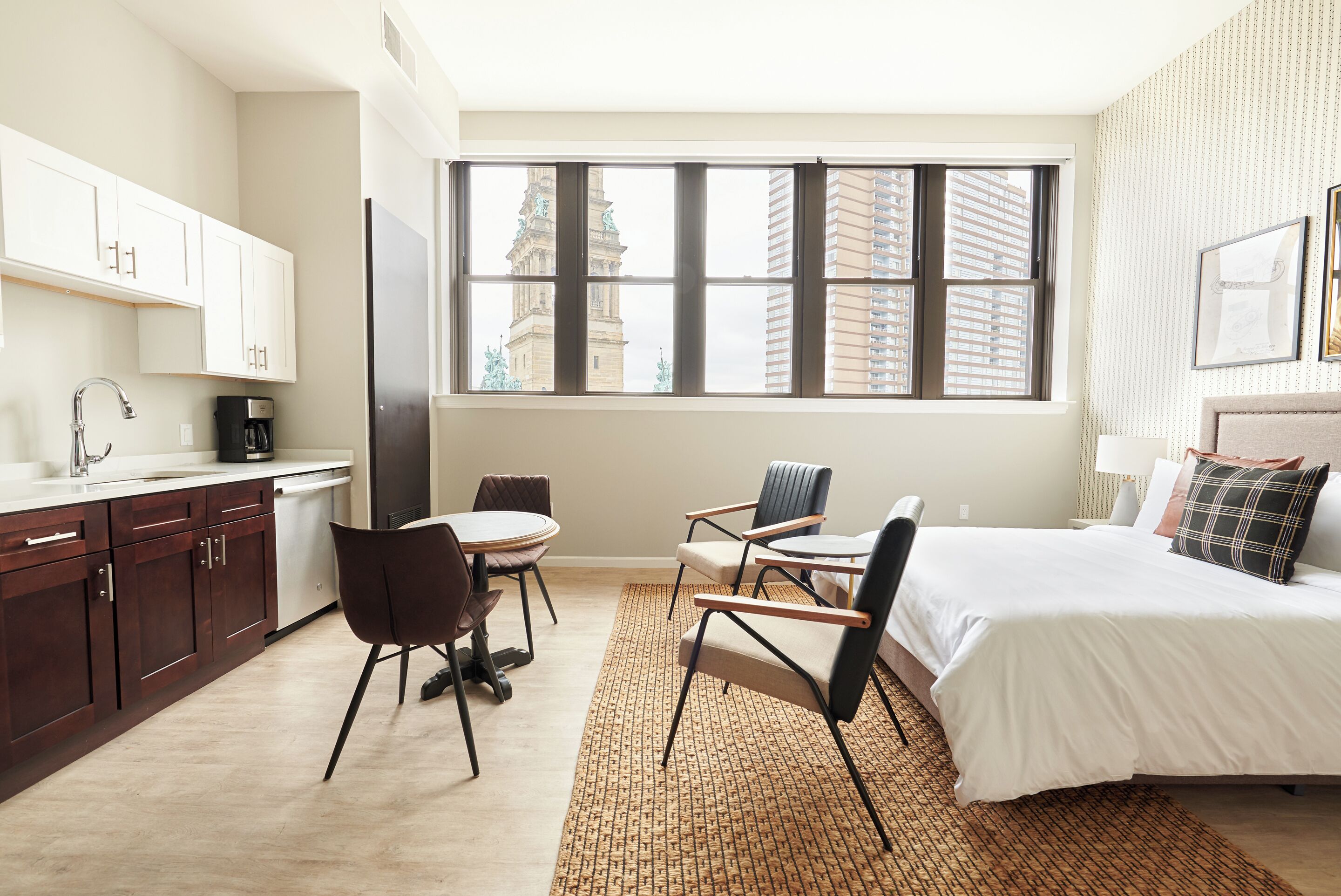 A modern hotel room with a large bed, a dining table with chairs, a kitchen area, and a view of a cityscape through large windows.