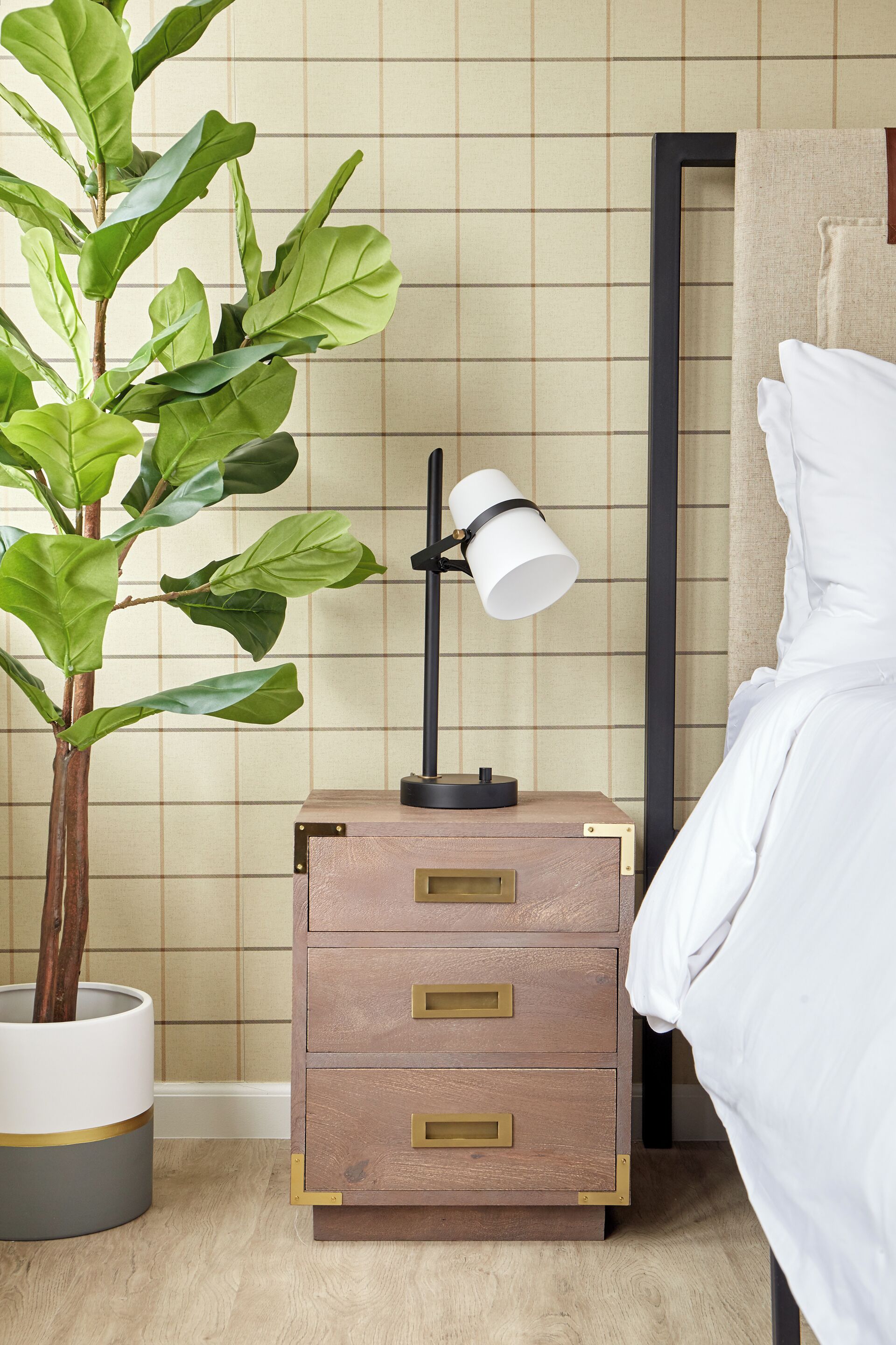 Una cama bien tendida con ropa blanca, una mesita de noche con lámpara y una planta grande en maceta.