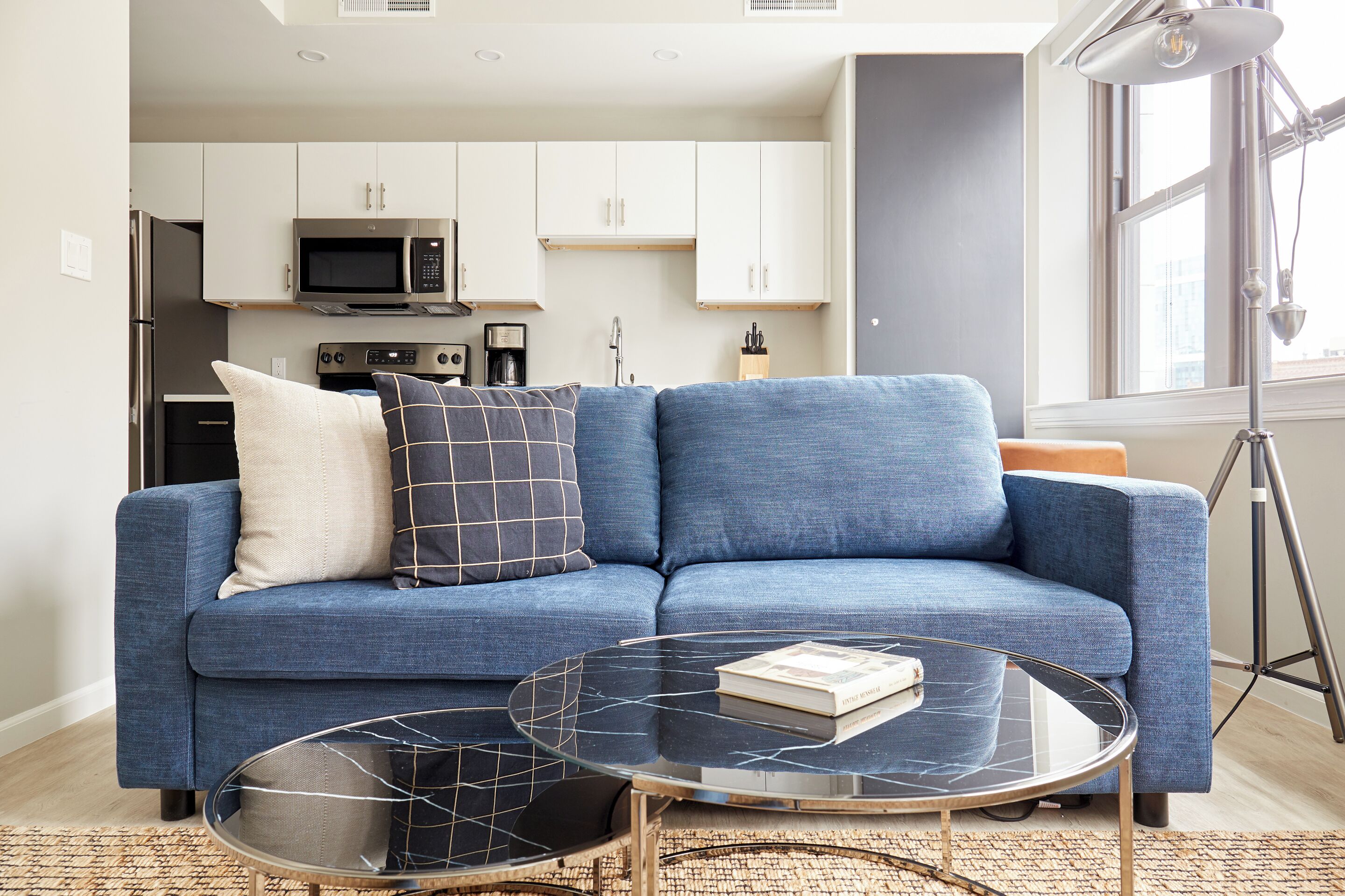 A modern living room with a blue sofa, a glass coffee table, and a kitchen in the background.