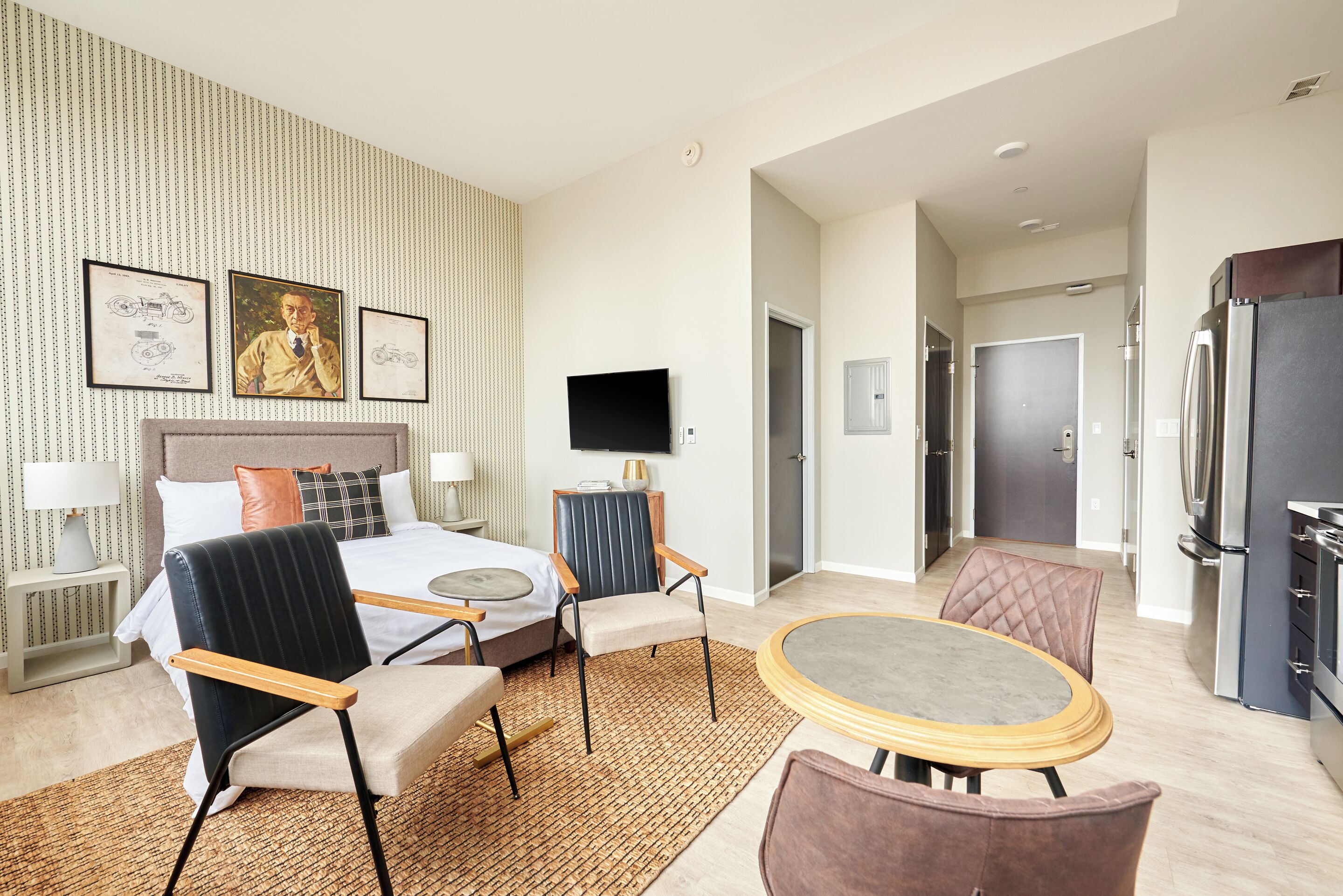 A hotel room with a bed, two chairs, a table, a TV, and a refrigerator.