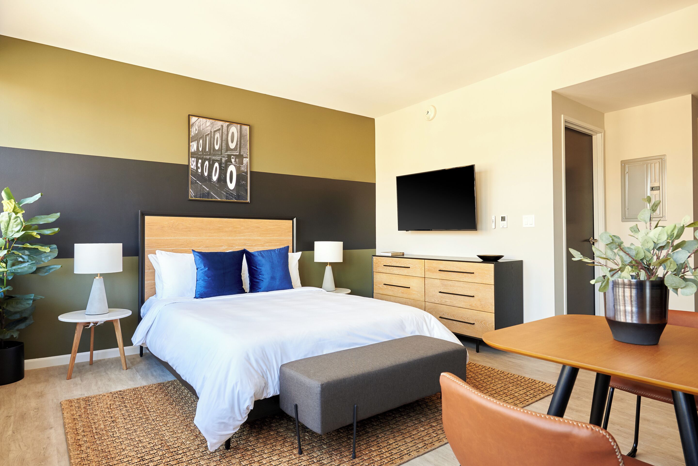 Ett modernt hotellrum med en stor säng, sängbord, en platt-TV, en byrå i trä och en matplats med bord och stolar.