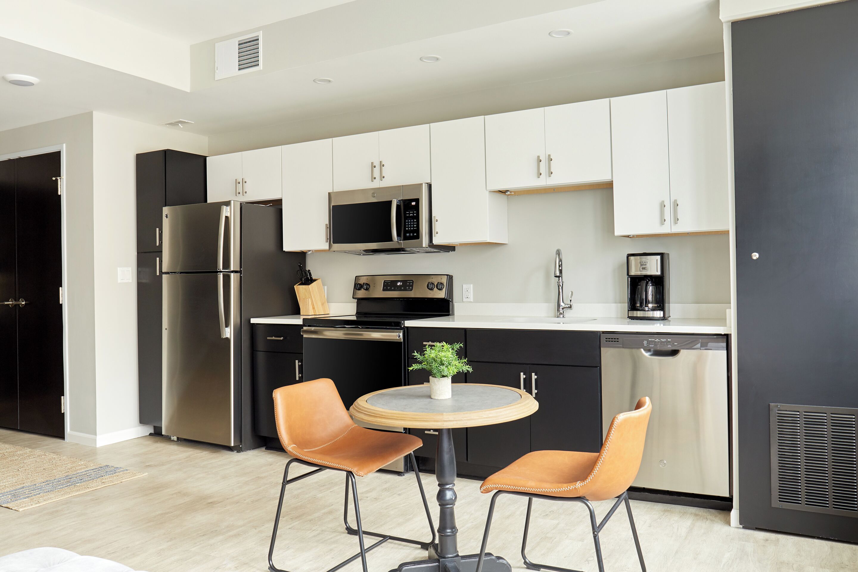 A modern kitchen with a stainless steel refrigerator, a microwave, a stove, and a dishwasher. There is a round table with two chairs and a small potted plant on it.