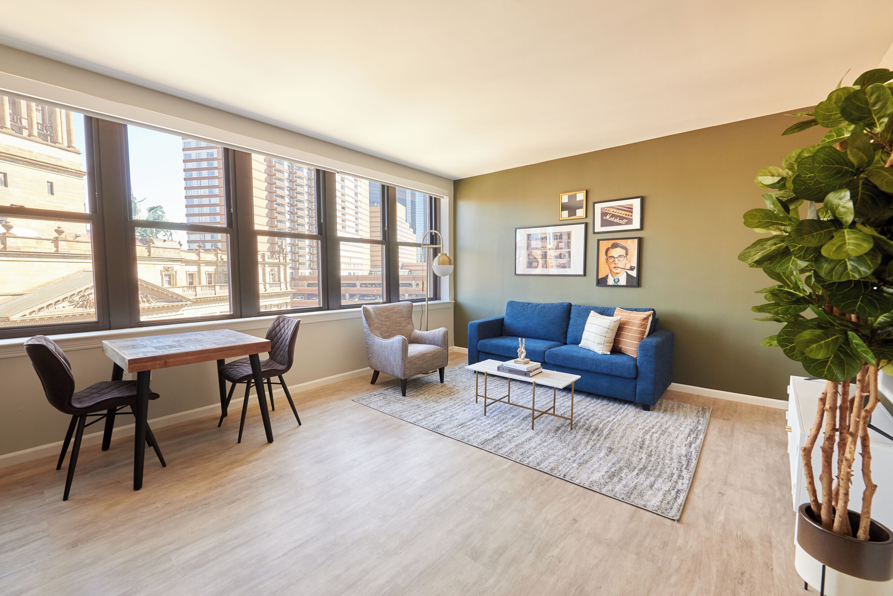A modern living room with a large window, a blue sofa, a dining table with chairs, and a potted plant.