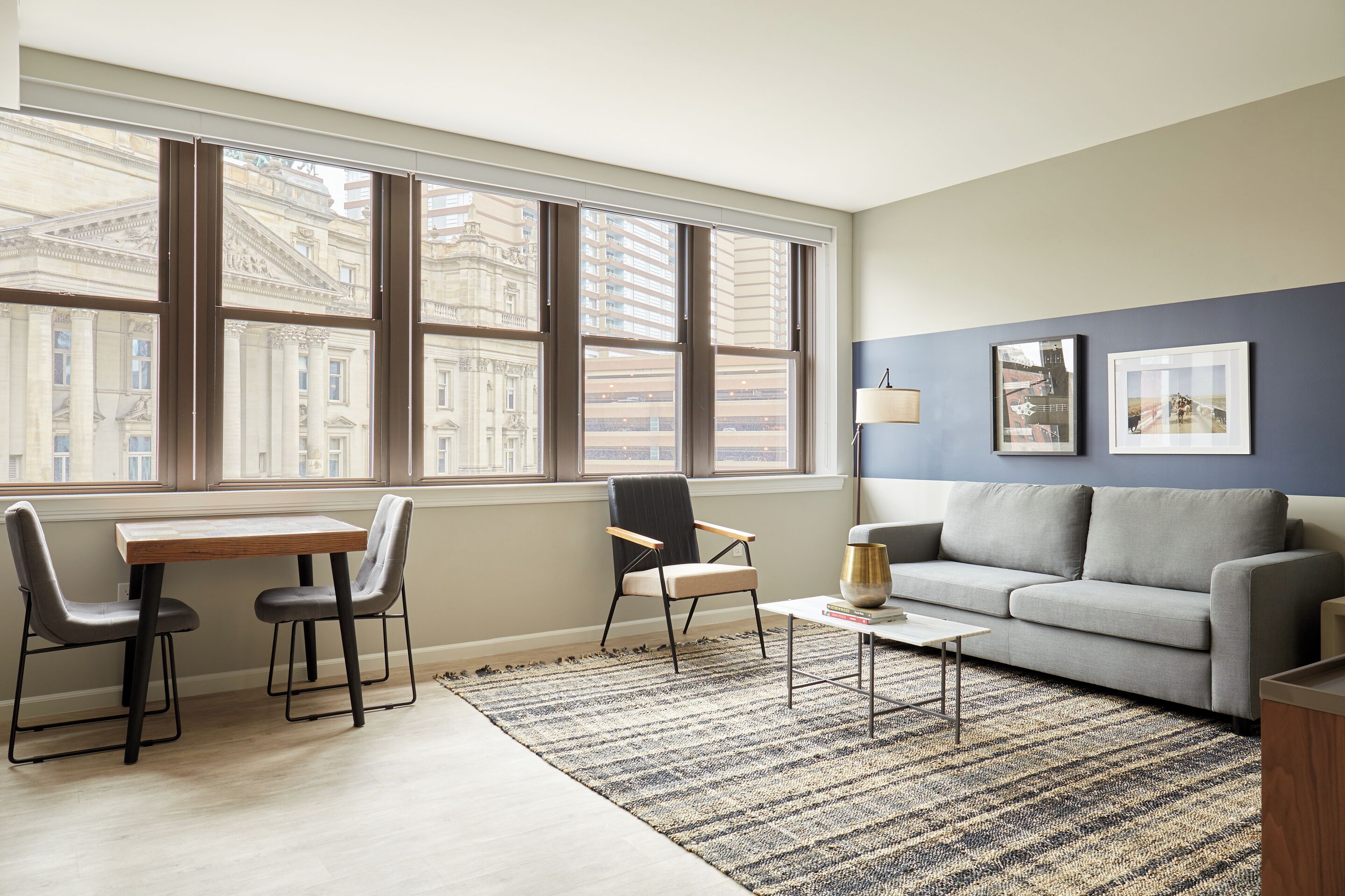 A modern living room with a large window, a grey sofa, two chairs, a coffee table, and framed pictures on the wall.