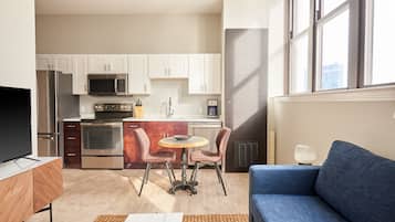 Une cuisine moderne équipée d’appareils électroménagers en acier inoxydable, un coin repas avec une table ronde et des chaises, et un canapé bleu dans un espace de vie lumineux doté de grandes fenêtres.