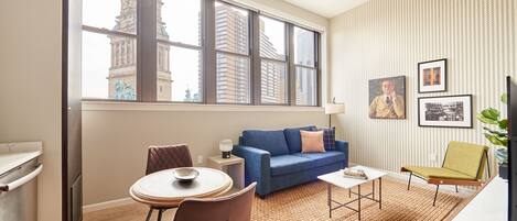 A modern living room with a large window, a blue sofa, a dining table, and a green chair.