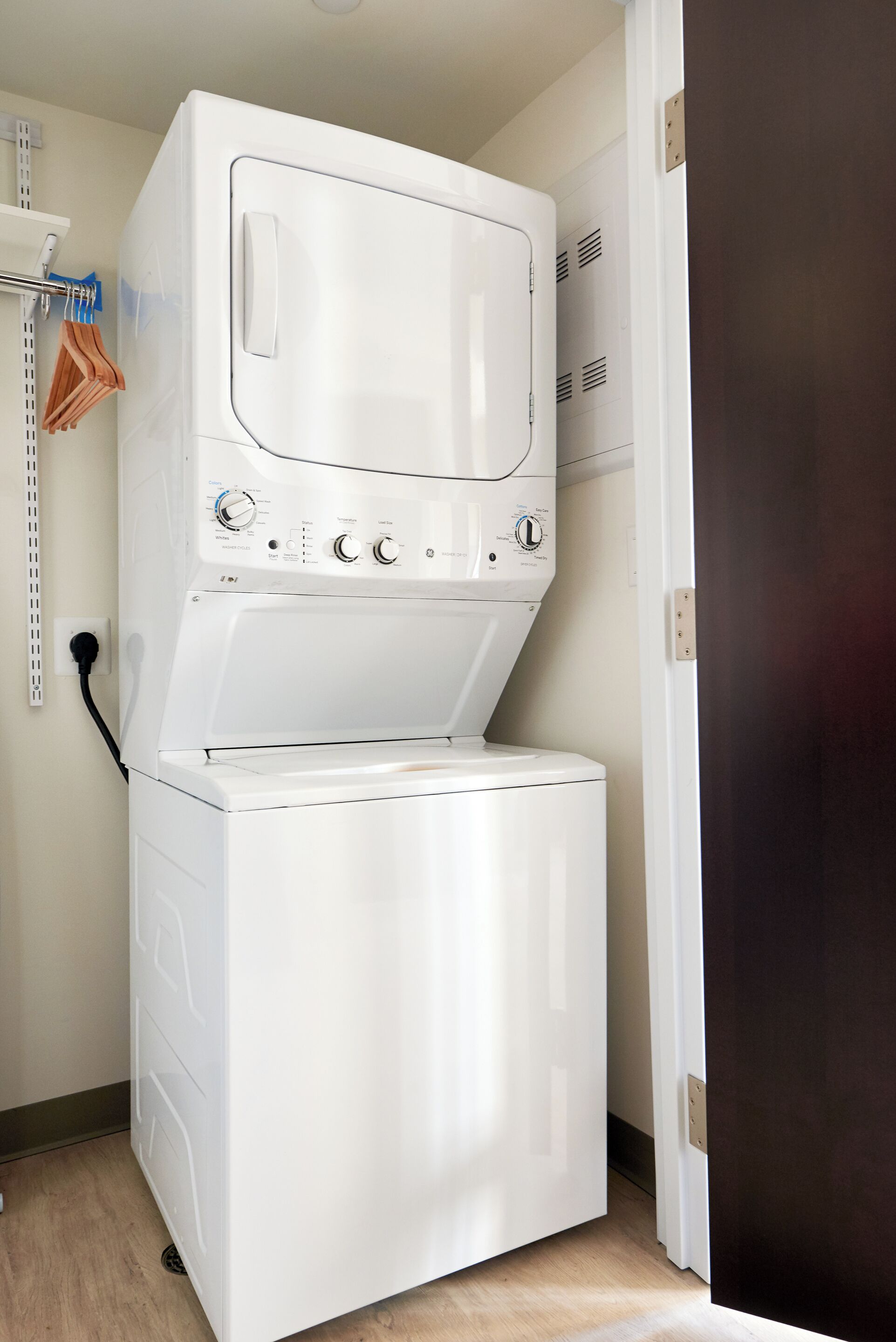 A compact laundry room with a stacked washer and dryer, a metal shelf with hangers, and a door to another room.