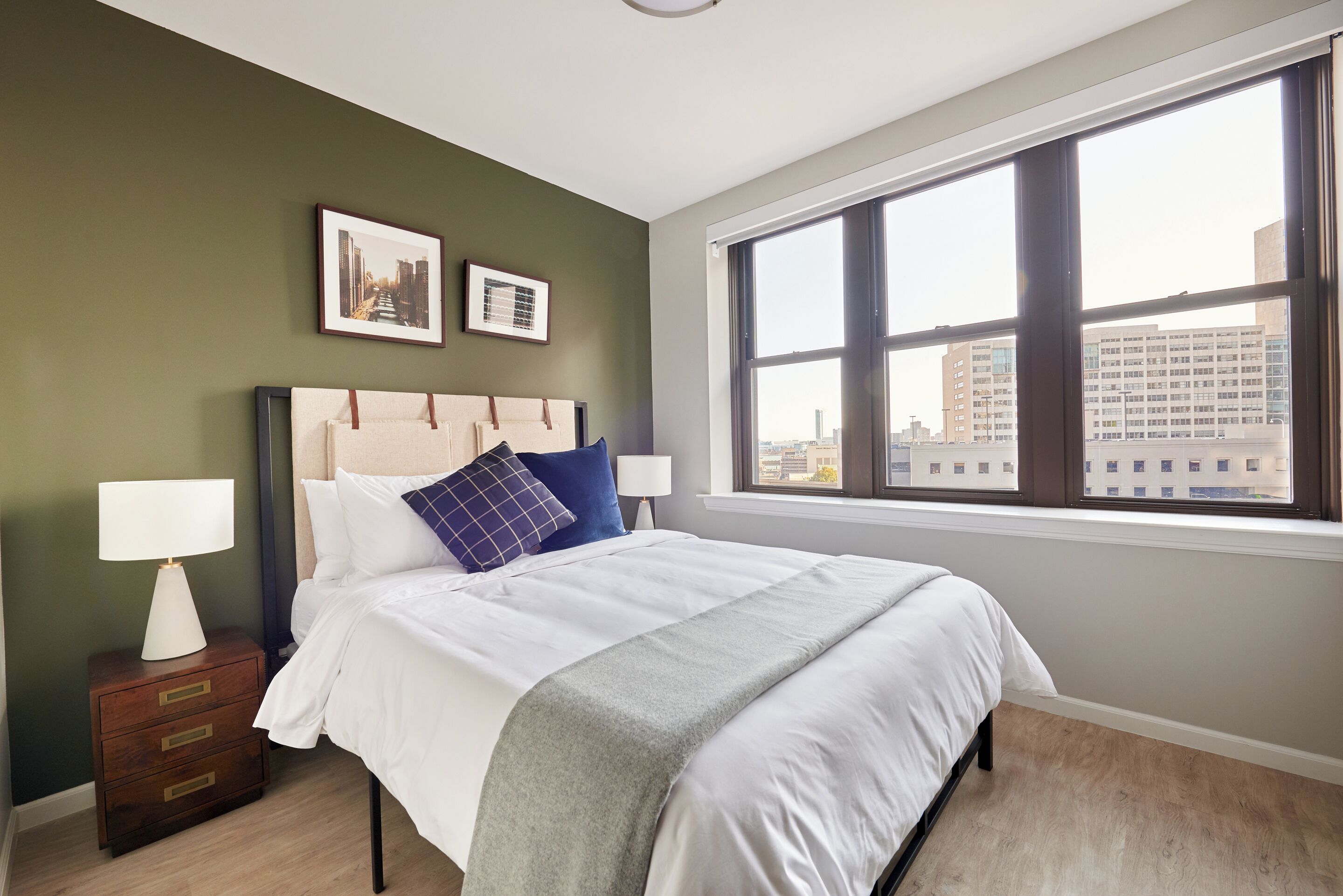A neatly made bed with a white comforter and a gray throw blanket, a nightstand with a lamp, and a window with a city view.