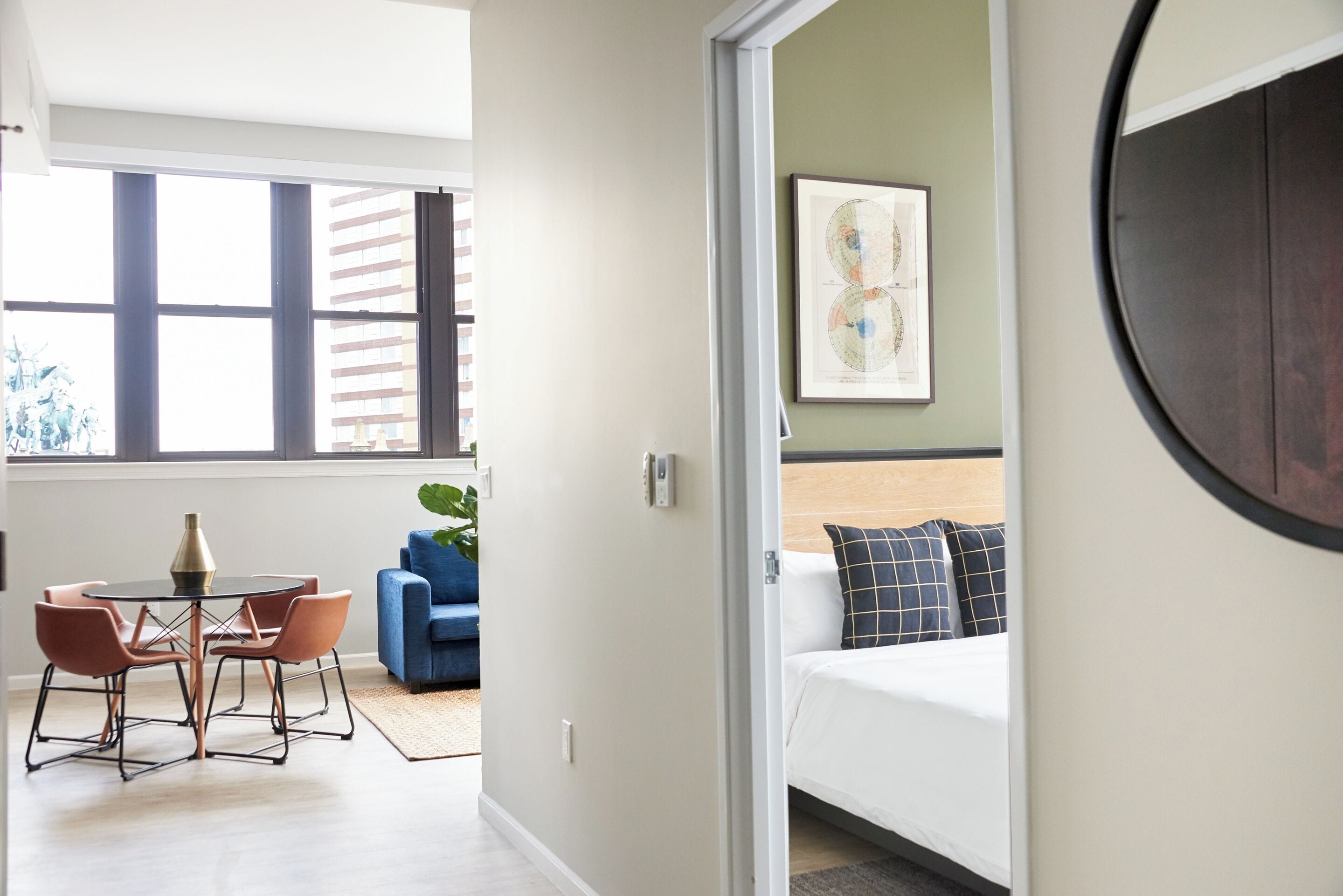 A modern hotel room with a bed, a dining area with a table and chairs, and a large window with blinds.