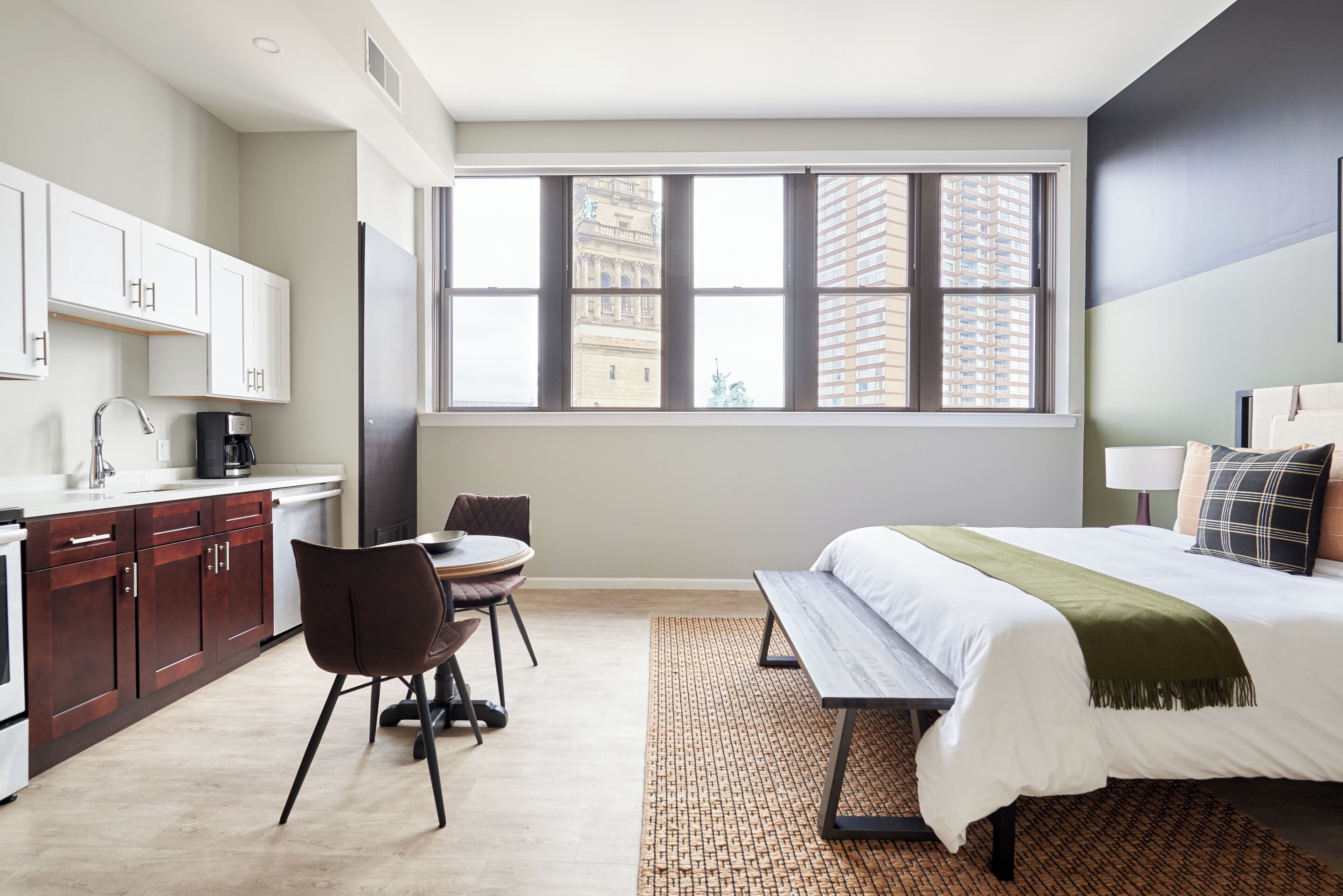 A modern hotel room with a large window, a bed, a desk, and a kitchenette.