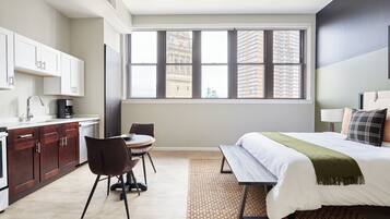 A modern hotel room with a large window, a bed, a desk, and a kitchenette.