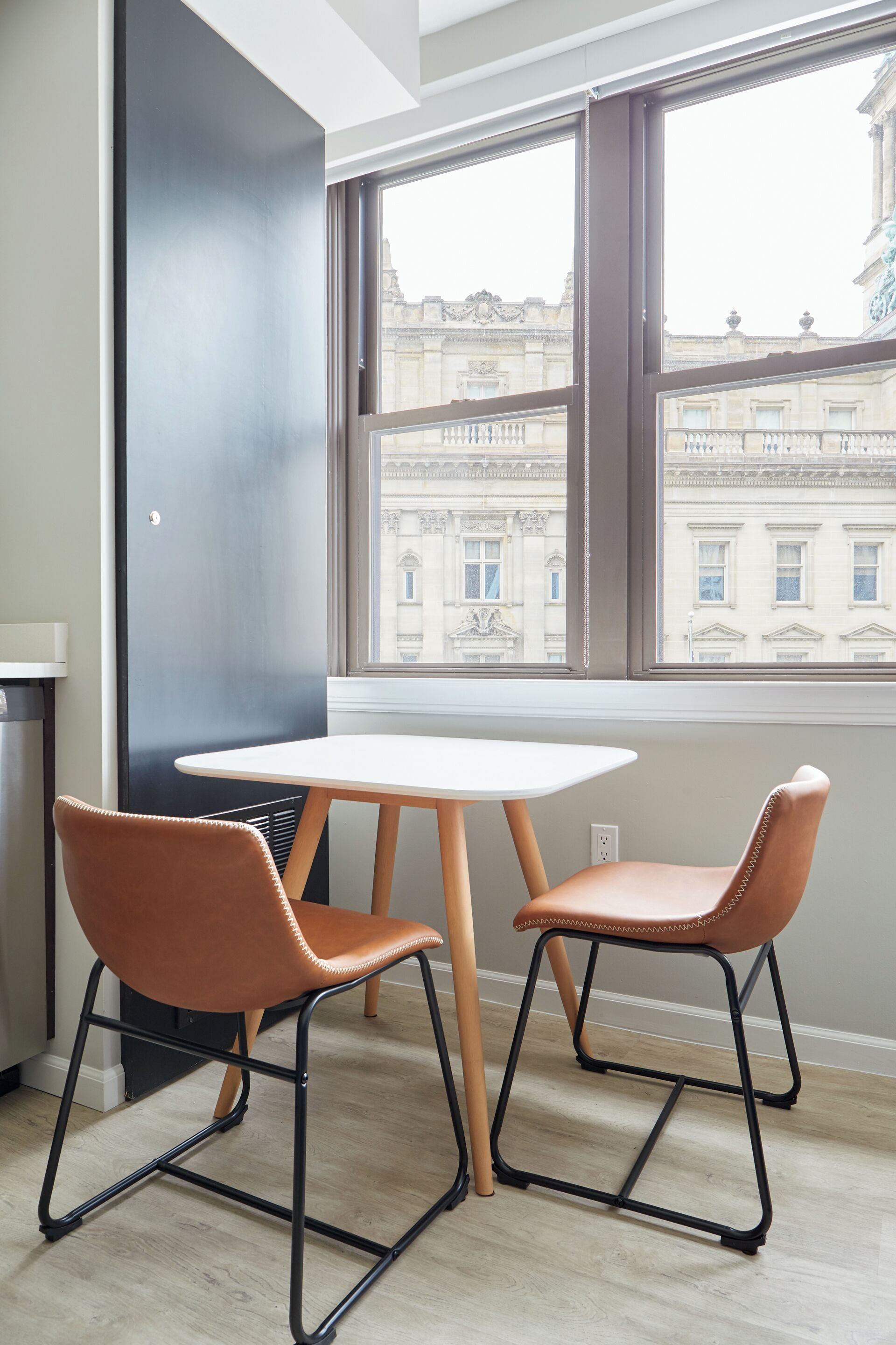 Une pièce avec une table blanche et deux chaises marron, une grande fenêtre donnant sur des bâtiments historiques et un mur d’accent noir.