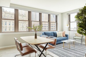 A modern living room with a dining table, blue sofa, and large windows offering city views.