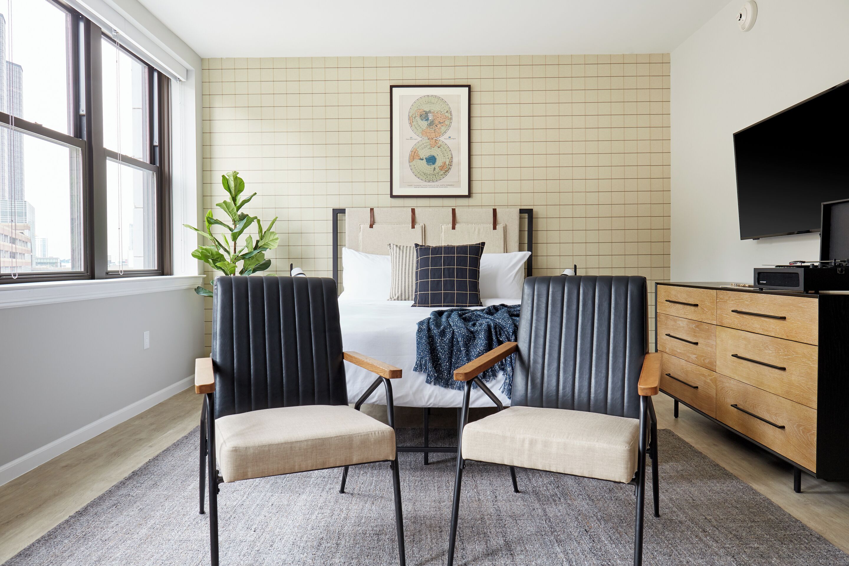 A bedroom with a bed, two chairs, a dresser, a television, and a window with a view of buildings.