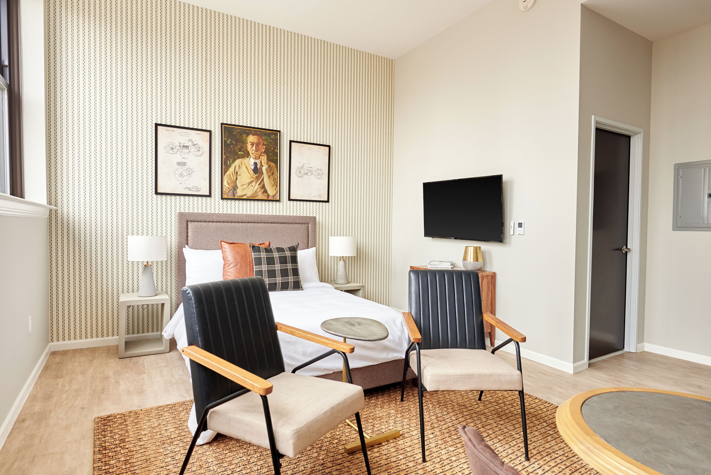 A hotel room with a bed, two chairs, a TV, and framed pictures on the wall.