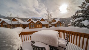A snow-covered deck with a chair and table, wooden cabins in the background, and a mountainous landscape. - Snake River 13 (Keystone)