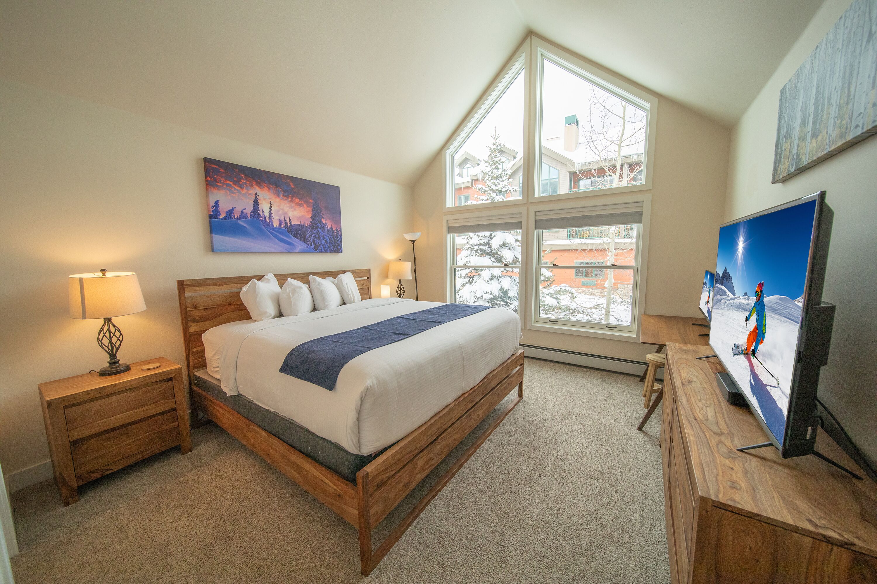A bedroom with a large bed, a nightstand, a dresser, a TV, and a window with a view of buildings.